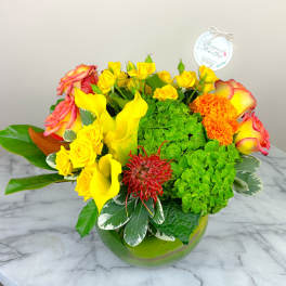 Bright mixed bouquet with yellow roses, calla lilies, and orange blooms in a green glass vase