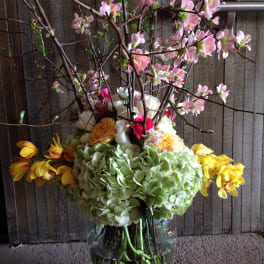 Tall floral arrangement with pink blossoms, hydrangeas, and yellow flowers in a glass vase