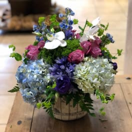 Round arrangement of blue hydrangeas, pink roses, purple blooms and white orchids in a ceramic pot