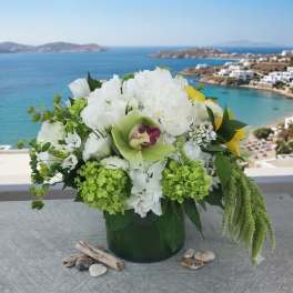 White and green floral arrangement in a dark vase with a seaside view behind it.