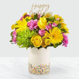 Bright yellow and pink bouquet in a white vase with a "Happy" topper.