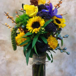 Tall bouquet of yellow roses, sunflowers, and purple flowers in a glass vase