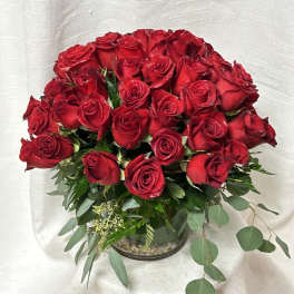 Round arrangement of many red roses in a clear glass vase with trailing eucalyptus.