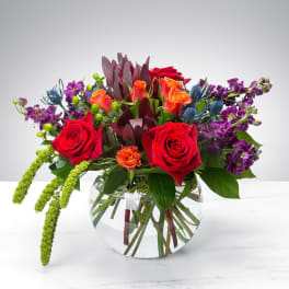 Mixed bouquet of red roses, orange blooms, and purple flowers in a glass vase