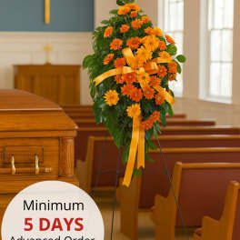 Standing spray of bright orange daisies with yellow bow on an easel beside a wood casket in a chapel.