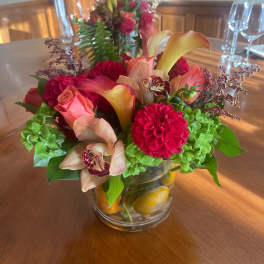 Low glass vase of red dahlias, peach roses, calla lilies, orchids, and green blooms on a wood table