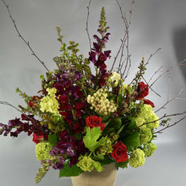 Tall floral arrangement with red roses, purple blooms, and green hydrangeas in a burlap-wrapped vase