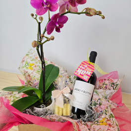 Pink orchid plant with a wine bottle and gift items in a wrapped basket