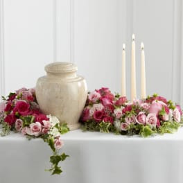 Cremation urn and three ivory candles encircled by pink and white rose flowers on a white table