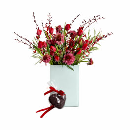 Red tulips and pink flowers in a tall glass vase with a wrapped chocolate heart