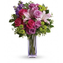 Tall arrangement of hot pink roses, lavender roses, lilies, and green button mums in a purple glass vase