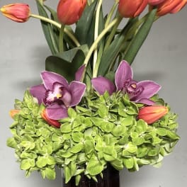 Orange tulips and purple orchids arranged in a tall glass vase