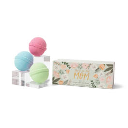Three pastel bath bombs beside a floral gift box labeled for mom.