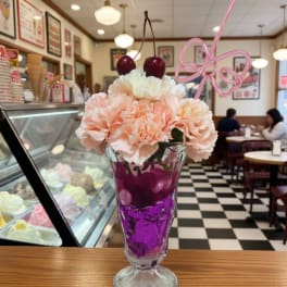 Peach carnations in a tall glass sundae dish with cherries and a pink straw bow