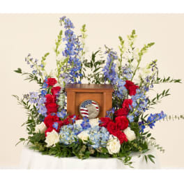 Floral tribute around a wooden urn with red, white, and blue flowers