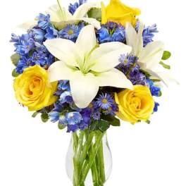 Clear vase of white lilies, yellow roses, and blue flowers in a rounded mixed bouquet