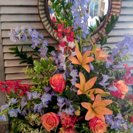 Tall arrangement of peach lilies, coral roses, blue delphinium and red orchids in a metal container