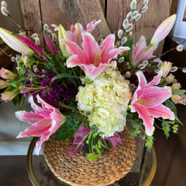 Arrangement of pink lilies, white hydrangea, and purple flowers in a vase with woven cover on a glass table.