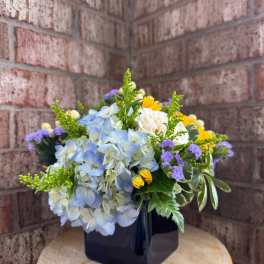 Blue hydrangea bouquet in a black square vase with yellow and purple accents