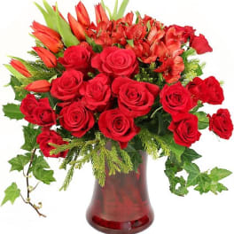 Arrangement of red roses, tulips, and alstroemeria in a red glass vase with trailing ivy.