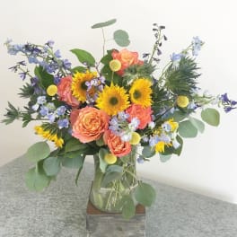 Mixed bouquet with orange roses, sunflowers, and blue flowers in a glass vase