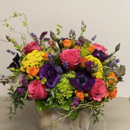 Vibrant arrangement of pink roses, purple blooms, orange flowers and green accents in a rustic ceramic container.