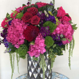Tall arrangement of hot pink hydrangeas, red roses, and purple blooms in a silver patterned vase