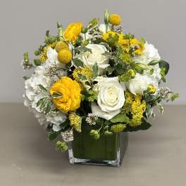 Rounded arrangement of white roses, hydrangeas, and yellow blooms in a clear square glass vase
