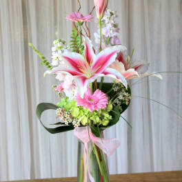 Pink lilies and gerbera daisies in a tall glass vase with a pink ribbon