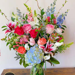 Tall mixed arrangement of pink roses, lilies, blue hydrangea and assorted blooms in a clear glass vase