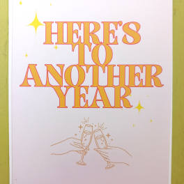 Greeting card with “Here’s to Another Year” text and hands clinking champagne glasses.