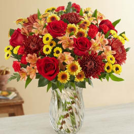 Mixed bouquet of red roses, yellow daisies, and orange flowers in a clear glass vase