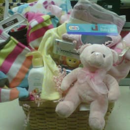 Baby gift basket with pink teddy bear, striped baby clothes, bibs, lotion, and blankets in a woven basket.