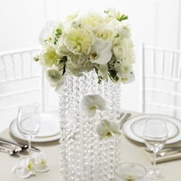 Tall white floral centerpiece with orchids and roses on a beaded glass vase on a formal table.