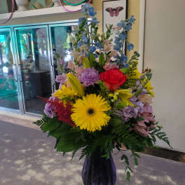 Tall mixed bouquet in a purple vase with blue, yellow, pink, and red flowers