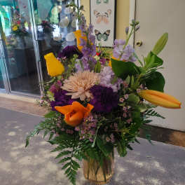 Mixed bouquet of yellow roses, orange calla lilies, and purple blooms in a gold vase
