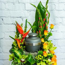 Cremation urn surrounded by orange roses, green orchids, and tropical flowers in a tall arrangement