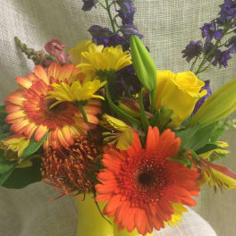 Bouquet of orange gerbera daisies, yellow roses, and purple flowers in a yellow vase