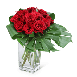 Red roses arranged in a clear square vase with large tropical leaves