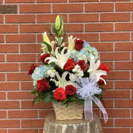 Basket arrangement of red roses, white lilies, and blue hydrangeas with a white bow