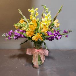 Mixed bouquet of yellow, orange, and purple flowers in a glass vase with a pink ribbon