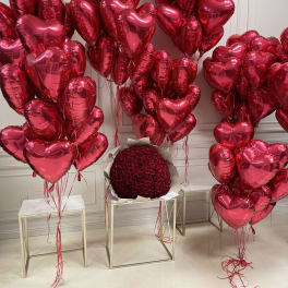 Clusters of red heart-shaped balloons surround a round bouquet of red roses.