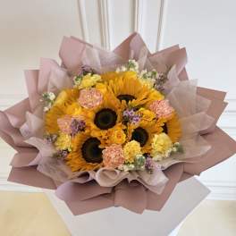 Bouquet of sunflowers, yellow roses, and pink carnations wrapped in blush paper