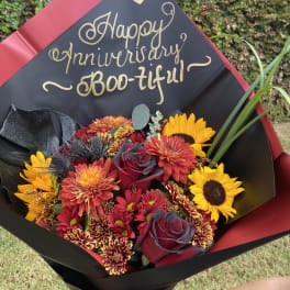 Bouquet of red, yellow, and orange flowers wrapped in black paper with a "Happy Anniversary" sign