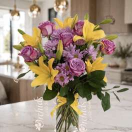 Bouquet of yellow lilies, purple roses, and lavender daisies in a glass vase