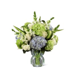 White roses, blue and green hydrangeas, and white snapdragons arranged in a clear glass vase