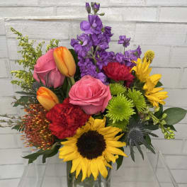 Mixed bouquet with roses, tulips, sunflowers, and purple blooms in a glass vase