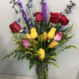 Tall bouquet of red and yellow roses with purple accents in a clear glass vase