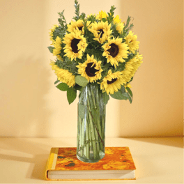 Tall bouquet of yellow sunflowers in a clear glass vase on a colorful book against a yellow background