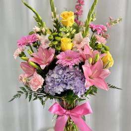 Mixed bouquet of pink lilies, yellow roses, and lavender hydrangea in a glass vase with a pink ribbon